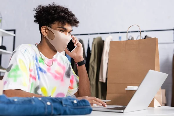 Propriétaire afro-américain de showroom en masque de protection parlant sur smartphone et utilisant un ordinateur portable près du sac à provisions — Photo de stock