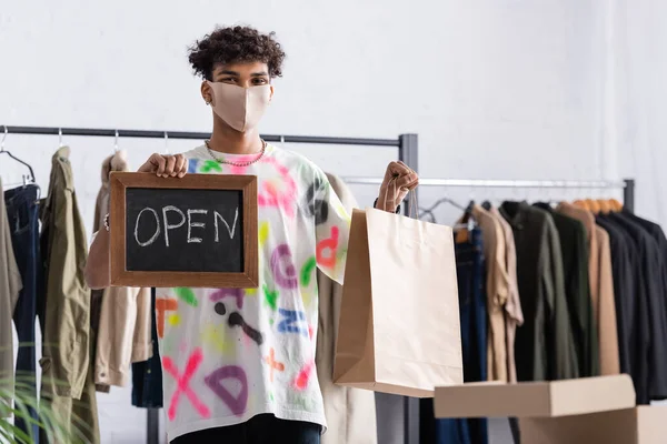 African american owner of showroom in protective mask holding chalkboard with open lettering and shopping bag — Stock Photo