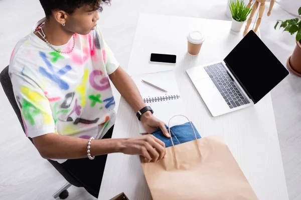 Laptop near african american owner of showroom putting clothes in shopping bag at table — Stock Photo