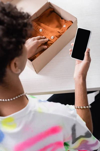 Вид зверху смартфона з порожнім екраном в руці власника афроамериканського шоуруму, який кладе одяг у коробку на розмитому тлі — стокове фото