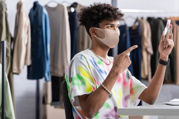 Propietario afroamericano del showroom en máscara protectora apuntando al desinfectante de la mano - foto de stock