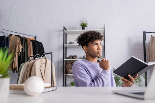 Pensive propriétaire afro-américain de showroom tenant stylo et ordinateur portable près de l'ordinateur portable sur le premier plan flou — Photo de stock