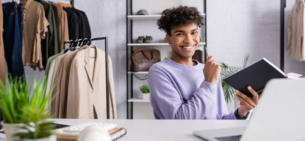 Propietario positivo afroamericano del showroom con la pluma y el cuaderno que mira la cámara cerca del ordenador portátil y de los pantalones, bandera - foto de stock