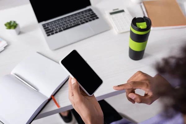 Vue grand angle d'un homme d'affaires afro-américain pointant vers un smartphone avec écran vide près d'un ordinateur portable et une tasse thermo sur fond flou — Photo de stock