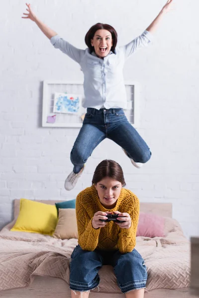 KIEW, UKRAINE - 20. NOVEMBER 2020: Teenagermädchen hält Steuerknüppel in der Nähe der erstaunten Mutter, die auf dem Bett springt — Stockfoto