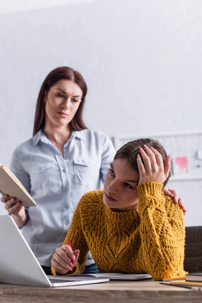 Bouleversé adolescent fille tenant stylo tout en faisant des devoirs près ordinateur portable et mère avec livre — Photo de stock