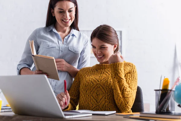 Excited teenage girl holding pen while doing homework near laptop and mother with book — Stock Photo