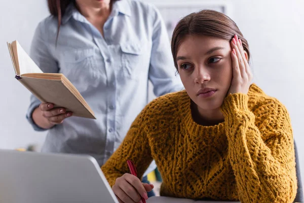 Gelangweilte Teenager mit Stift bei den Hausaufgaben in der Nähe von Laptop und Mutter mit Buch — Stockfoto