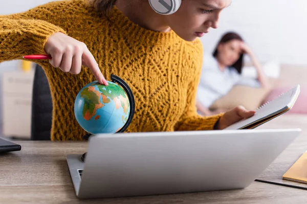 Teenager-Mädchen mit drahtlosen Kopfhörern zeigt auf Globus, während sie auf Notizbuch nahe verschwommener Mutter im Hintergrund schaut — Stockfoto