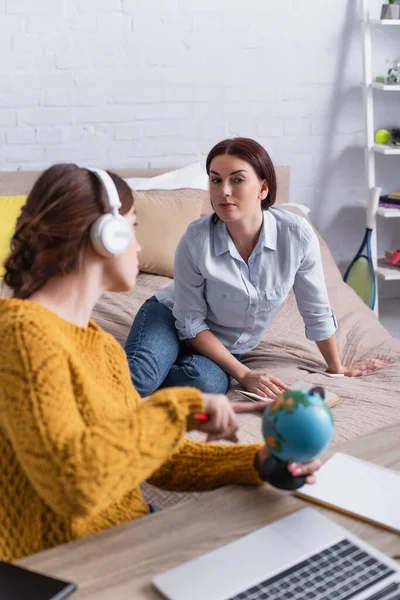Blurred teenage girl in wireless headphones pointing at globe while looking at mother — Stock Photo
