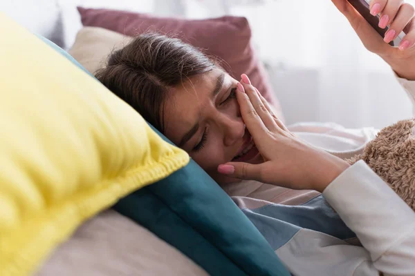 Frustrado adolescente cubriendo la boca mientras llora y sostiene el teléfono inteligente en el dormitorio - foto de stock