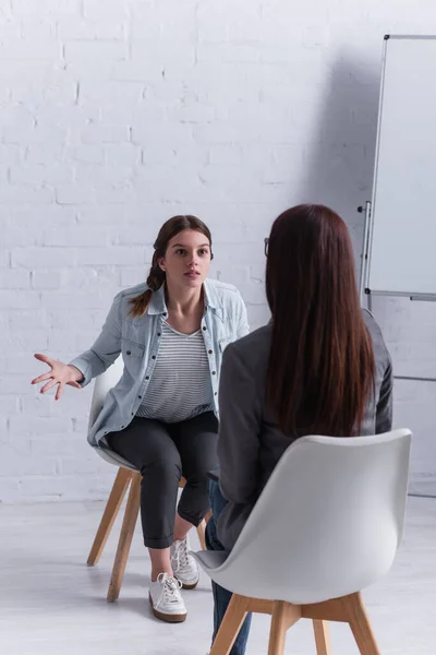 Disgustada adolescente sentada y haciendo gestos mientras mira al psicólogo en primer plano borroso - foto de stock