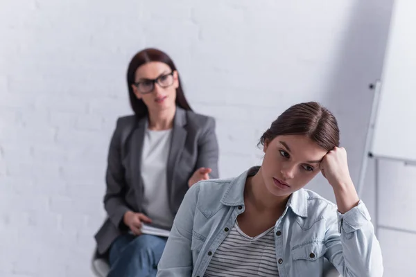 Worried teenage girl looking away with psychologist sitting behind on blurred background — Stock Photo