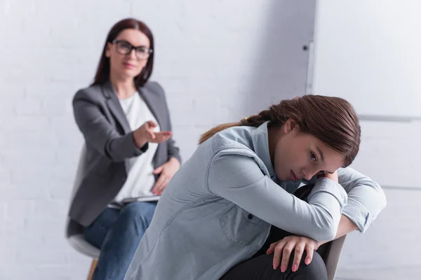 Frustrated teenage girl looking away with psychologist sitting behind on blurred background — Stock Photo
