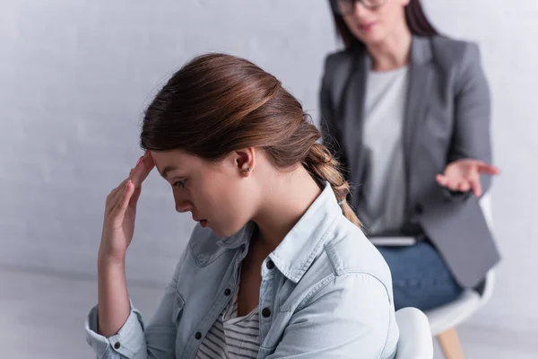 Deprimida adolescente mirando hacia otro lado cerca psicólogo sentado detrás en fondo borroso - foto de stock