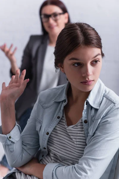 Depressed teenage girl looking away and gesturing near psychologist sitting behind on blurred background — Stock Photo