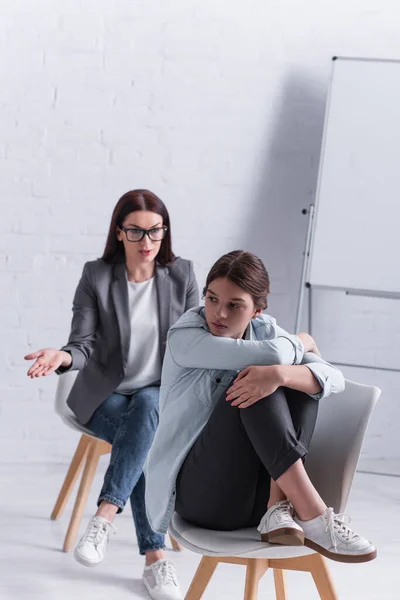 Depressed teenage girl looking away and sitting near psychologist in clinic — Stock Photo