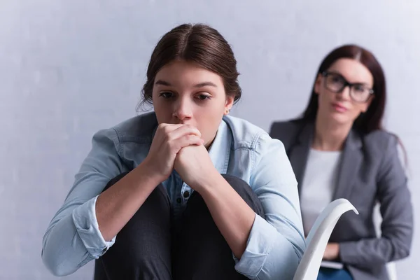 Depressed teenage girl with clenched hands near psychologist sitting behind on blurred background — Stock Photo