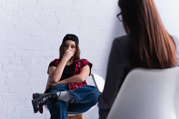 Sad teenage patient in cap sitting and covering mouth near psychologist on blurred foreground — Stock Photo