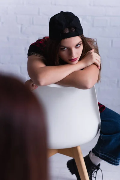 Upset teenage patient in cap sitting and looking at psychologist on blurred foreground — Stock Photo