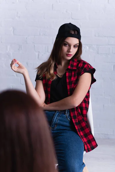 Skeptical teenage patient in cap looking at psychologist on blurred foreground — Stock Photo