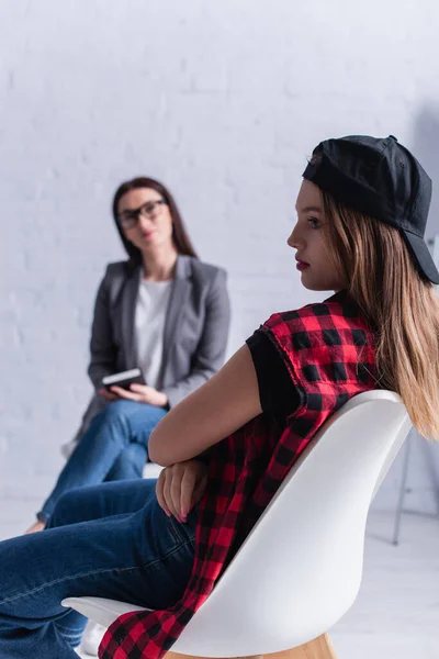Sad teenage girl in cap looking away during therapy with psychologist on blurred foreground — Stock Photo