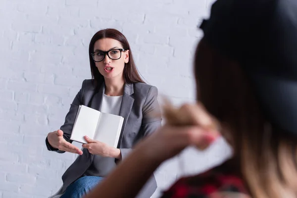 Psychologist in glasses holding blank notebook and gesturing while looking at teenage patient on blurred foreground — Stock Photo