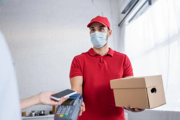 Courier musulman dans le masque médical tenant terminal de paiement près de la femme avec smartphone au premier plan flou — Photo de stock