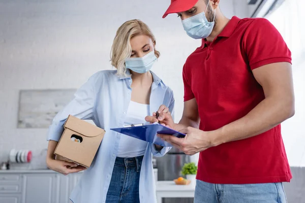 Muslim delivery man writing on clipboard near woman in medical mask holding parcel — Stock Photo
