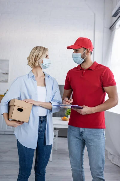 Arabian delivery man looking at blonde woman holding parcel at home — Stock Photo