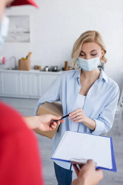 Woman in medical mask taking pen from postman on blurred foreground — Stock Photo