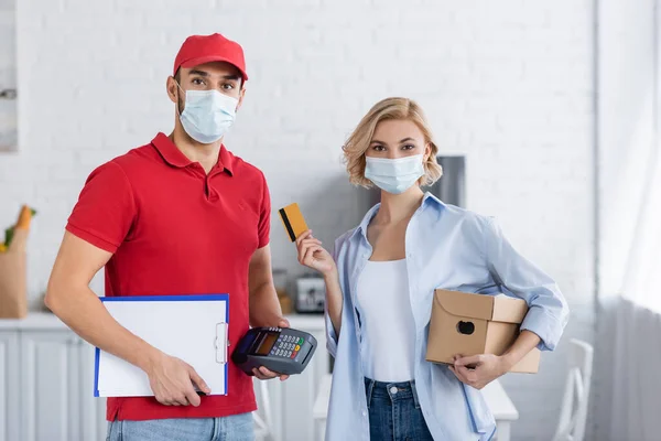 Arabe livraison homme et femme blonde dans des masques médicaux regardant la caméra à la maison — Photo de stock