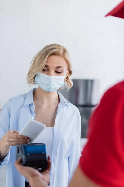 Repartidor en borroso primer plano tenencia terminal de pago cerca de la mujer en máscara médica, borrosa primer plano - foto de stock