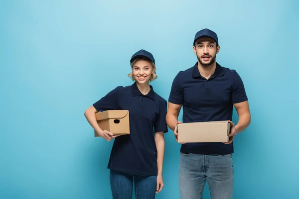 Mensajeros multiculturales felices sosteniendo cajas de cartón mientras mira la cámara en azul - foto de stock