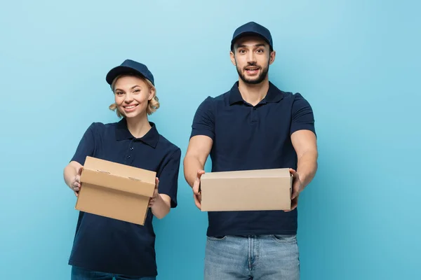 Positive interracial couriers showing cardboard boxes while looking at camera isolated on blue — Stock Photo