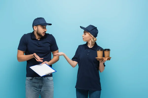 Mensajero con bebida para llevar apuntando a confuso colega árabe con portapapeles vacío aislado en azul - foto de stock
