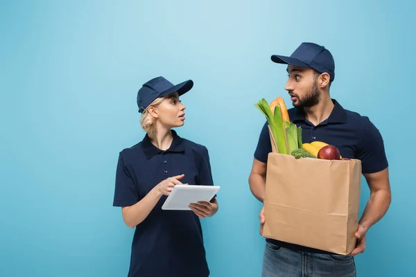 Young courier pointing at digital tablet near surprised arabian colleague with food isolated on blue — Stock Photo