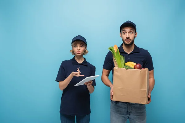 Correio chateado apontando para tablet digital perto de correio árabe surpreso segurando saco de papel com alimentos isolados em azul — Fotografia de Stock