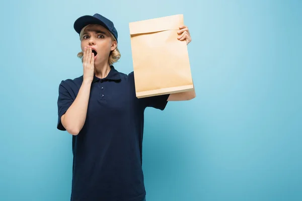 Impactado mensajero que cubre la boca con la mano mientras sostiene la bolsa de papel en azul - foto de stock