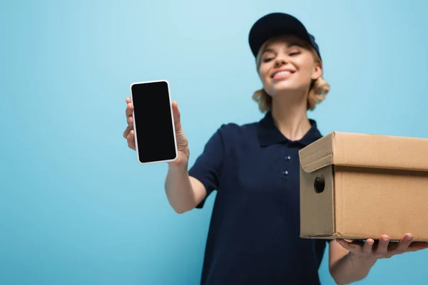 Smiling courier showing smartphone with blank screen isolated on blue, blurred background — Stock Photo