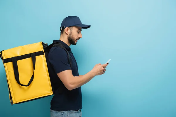 Side view of arabian delivery man messaging on mobile phone while carrying thermo bag on blue — Stock Photo