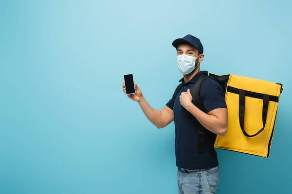 Мусульманский курьер в медицинской маске показывает мобильный телефон с пустым экраном на синем — стоковое фото