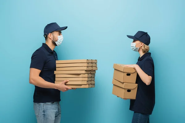 Side view of interracial couriers in medical masks holding packages on blue — Stock Photo