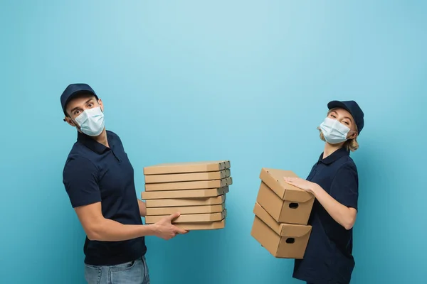 Multicultural couriers in protective masks, with pizza boxes and packages on blue — Stock Photo