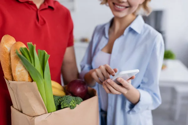 Teilbild eines Lieferanten mit frischen Lebensmitteln in Papiertüte neben glücklicher Frau, die auf verschwommenem Hintergrund auf Smartphone zeigt — Stockfoto