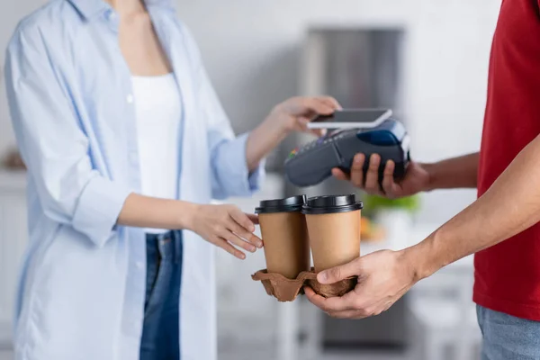 Vista parziale di donna che tiene smartphone vicino all'uomo di consegna con caffè da andare e terminale di pagamento su sfondo sfocato — Foto stock