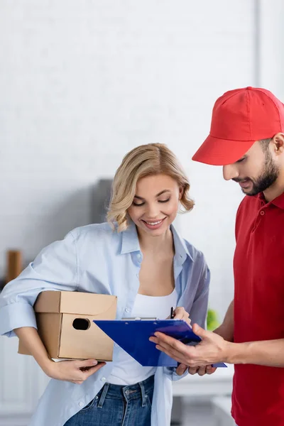 Smiling woman signing order on clipboard near arabian delivery man — Stock Photo