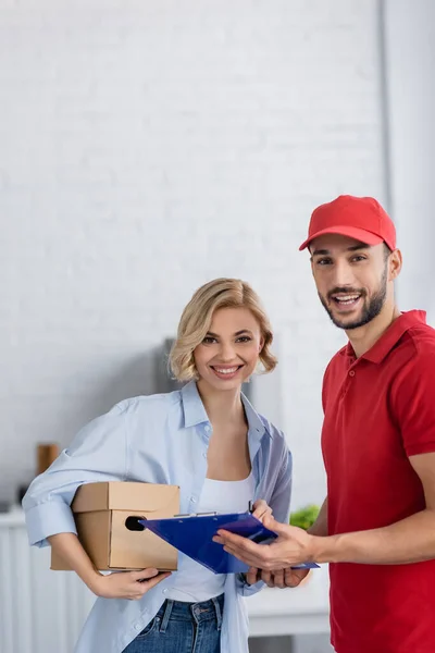 Happy arabian delivery man and blonde woman looking at camera at home — Stock Photo