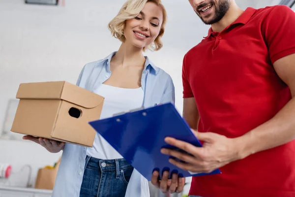 Low angle view of delivery man holding clipboard near blonde woman, blurred foreground — Stock Photo