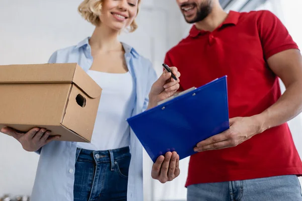 Cropped view of courier holding clipboard near smiling woman with parcel, blurred background — Stock Photo